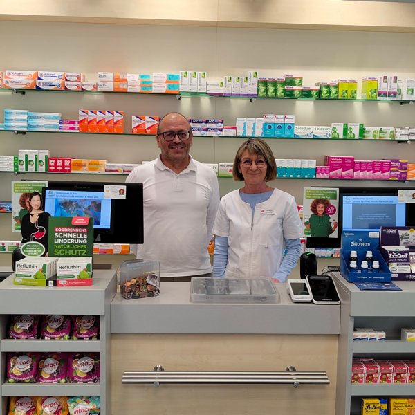 Team der Lonetal Apotheke OHG an der Kasse Zwei Mitarbeitende der Lonetal Apotheke OHG stehen hinter dem Kassentresen. Im Hintergrund sind Arzneimittelregale mit farblich sortierten Produkten zu sehen. Auf dem Tresen stehen Displays mit Gesundheitsprodukten und zwei Monitore.